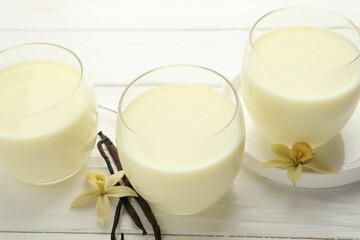 Delicious vanilla milk in glasses, pods and flowers on white wooden table