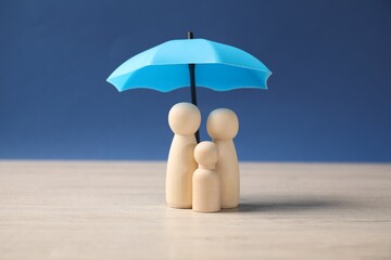 Human figures under umbrella on light wooden table against blue background. Insurance concept