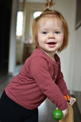 Naklejka premium Red hair baby girl sitting on wooden tricycle at home. Toddler girl riding a three wheel bike indoors.