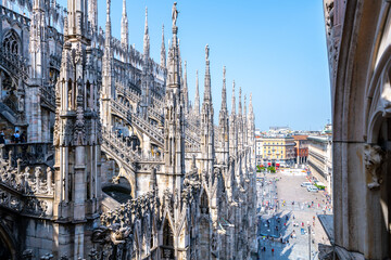 Fototapeta premium The stunning architecture of the Duomo di Milano is visible from the rooftop, showcasing intricate spires and a vibrant urban square bustling with visitors under a clear blue sky.