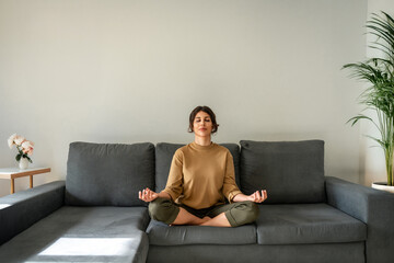 Relaxed young indian woman sitting on couch, meditating with closed eyes in lotus pose. Peaceful...