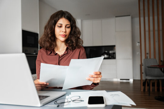 Focused latin hispanic young business woman working on laptop computer reading financial document report at home office. Accountant entrepreneur manager businesswoman doing paperwork using pc remotely