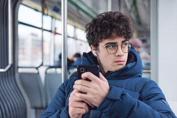 Teen boy in blue jacket rides public transport, listening to music on earbuds. He looks at his phone. Blurred background reveals passengers and bus/tram interior. urban commute moment. Сopyspace © Светлана Мищенко