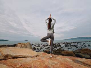 Young woman practicing yoga on a rocky beach, showing tranquility and focus, wearing a gray top and...