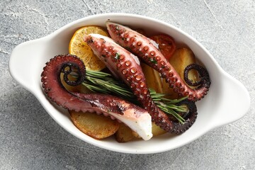 Fried octopus with vegetables, lemon and rosemary in baking dish on grey textured table, top view