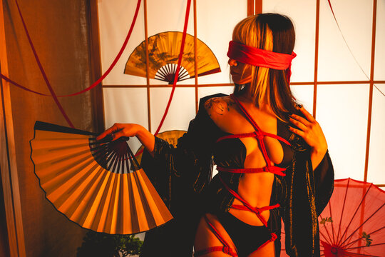 Beautiful young woman with blonde hair, black kimono and panties, and red shibari ties and ropes, red blindfold, holding a paper fan in a warm japanese room