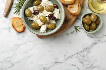 Marinated olives with feta cheese, bread pieces, oil and rosemary on white marble table, flat lay....