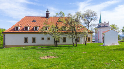 Fototapeta premium A serene view of the Skalka Baroque complex near Mnisek pod Brdy, showcasing the historic architecture, lush green lawn, and clear blue sky on a sunny day.