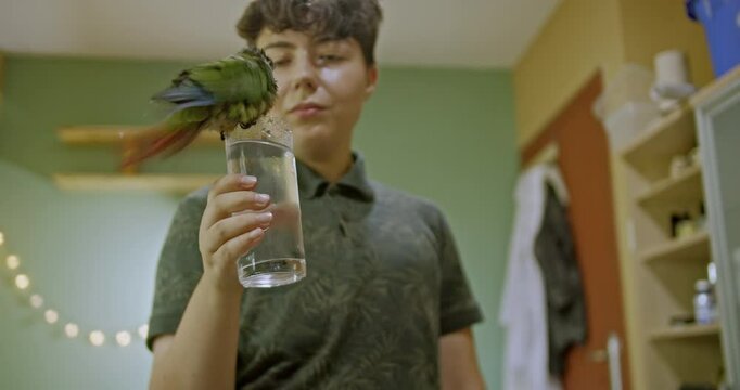 A girl offers a glass of water to her pet parrot in her bedroom, watching as the bird drinks and splashes happily, enjoying the cool refreshment.