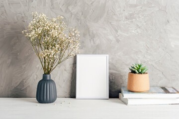 Blank frame with a vase of white gypsophila flowers and potted succulent on the stack of books. Mockup