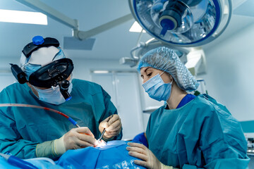 Surgeons in a sterile room. Two surgeons in blue scrubs, focused on surgery amid bright lights and...