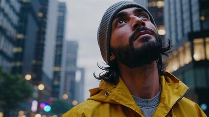 Young traveler wearing bright yellow raincoat standing amid urban skyscrapers, gazing upward with architectural wonder during evening cityscape exploration