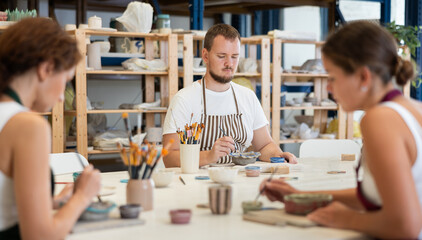 In pottery workshop, two girls and guy are sitting at table, busy coloring products made of raw clay. Amateurs are busy painting clay plates at master class