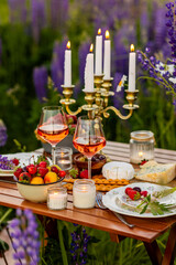 Two glasses of rose wine, flowers in a vase, silverware, fruits. Sunset, golden hour, atmospheric candlelight. Romantic table decor for a loving couple on blooming meadow with purple lupines