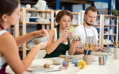 Three craftspersons in striped aprons are busy molding clay products. They are sitting near work table, making plate out of raw clay. Working together in pottery workshop.