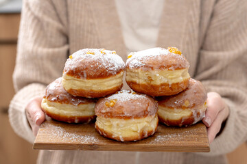 A cozy home setting with fresh Polish doughnuts (paczki) being sprinkled with powdered sugar. Soft, golden-brown pastries with icing and candied orange peel, perfect for Fat Thursday celebrations.