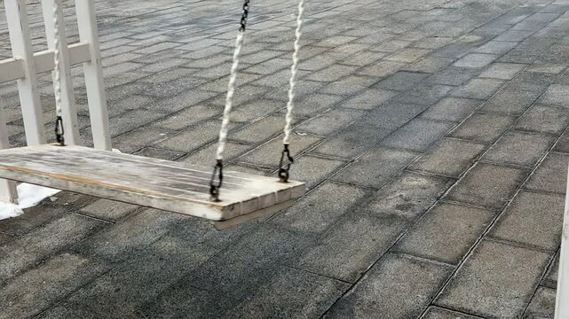 An old swing sways softly in an empty courtyard, surrounded by stone tiles. The atmosphere is calm and serene as the cool breeze encourages movement.