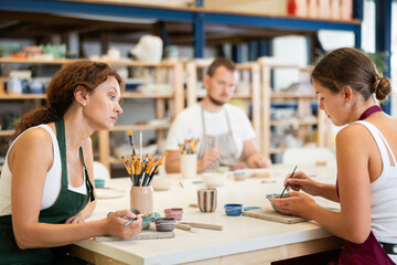 In pottery workshop, two girls and guy are sitting at table, busy coloring products made of raw clay. Amateurs are busy painting clay plates at master class
