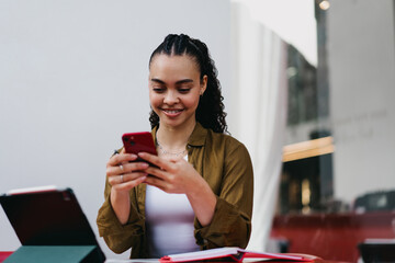 Smiling Hispanic woman using smartphone at outdoor café, engaging with social media or messaging. female Digital nomad enjoying remote work flexibility, connected lifestyle, and online communication