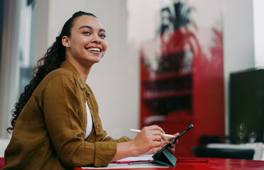 Smiling young Latina freelancer working remotely at cozy café, using tablet and stylus for...