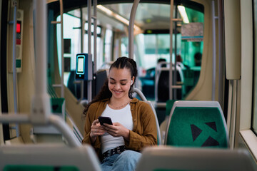 Young Latina woman sitting comfortably in tram, using smartphone while wearing wireless earbuds....