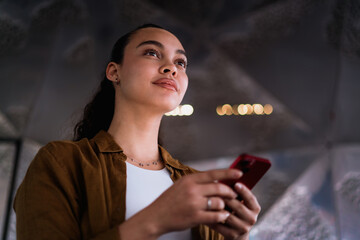 Attractive Latina woman looking up while holding a smartphone, surrounded by a futuristic metallic...