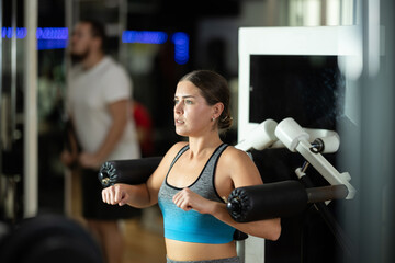 Fototapeta premium Woman in a sports top performs exercises for her arms in a simulator. European woman does shoulder abductions for the deltoid muscles. Exercises for strength and relief of the arms