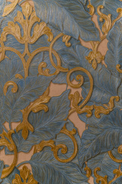 A close-up of an elegant wallpaper or textile with intricate blue and gold floral patterns. The raised textures and swirling details create a luxurious and ornamental feel.