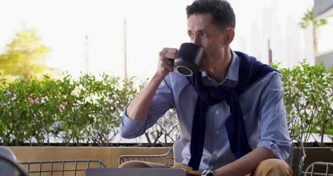 A respectable man is drinking coffee sitting on the cafe terrace in the city center. An open laptop on the table in front of him