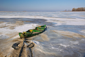 Old abandoned green and yellow wooden fishing boat stuck in the melting ice of a frozen lake at the...
