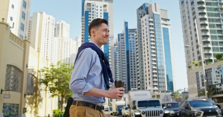 A respectable man crosses the road at a pedestrian crossing. He holds a disposable cup of coffee and a laptop in his hands.