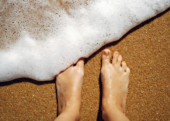 Woman looking down on feet standing on sand and crashing wave in Portugal beach