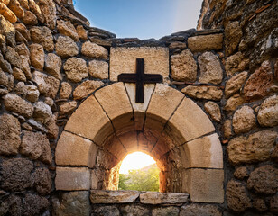 A historic stone archway with a black cross at the top frames a beautiful sunset in the background.