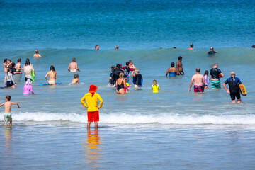 A lifeguard in a bright color uniform stands at the beach, watching numerous people or beachgoers...
