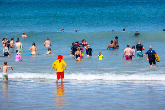 A lifeguard in a bright color uniform stands at the beach, watching numerous people or beachgoers swim and enjoying the waves on a summer day at Great Ocean Road. Torquay, VIC Australia. - Powered by Adobe