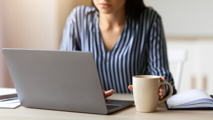 Fototapeta premium Businesswoman's Lifestyle. Unrecognizable Woman Working On Laptop Sitting At Workplace In Office, Focus On Coffee Cup. Cropped, Shallow Depth
