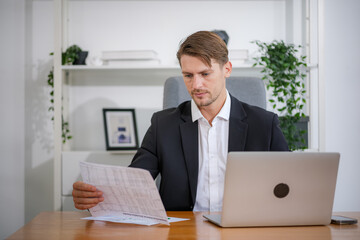 The male executive is sitting at her desk reading her financial report and comparing the results on her desktop.