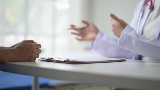 A doctor and patient are sitting at a desk with a clipboard in front of them. The patient is looking at the doctor with a concerned expression. The doctor is holding a pen