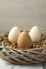 Egg-shaped candles in nest on white marble table against light gray background, closeup. Easter decor