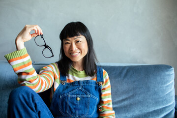 Portrait of a smiling happy Asian woman with glasses sitting on sofa in colorful outfit