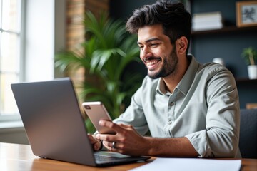 Fototapeta premium Young South Asian Male Smiling While Using Smartphone and Laptop in a Bright Home Office Environment with Indoor Plants
