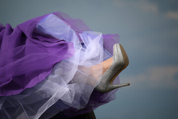 A silver colored stiletto covered in sparkly gems is raised in the air by an unrecognizable woman wearing a purple evening dress with crinoline.