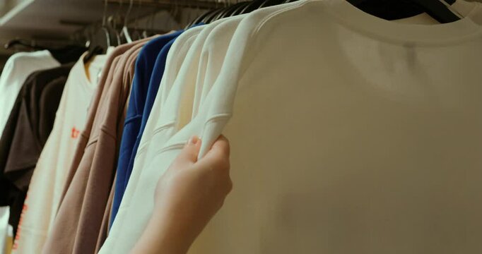 White hoodie gripped in hands, carefully inspecting fabric texture amid surrounding clothing displays, capturing detailed retail shopping moment