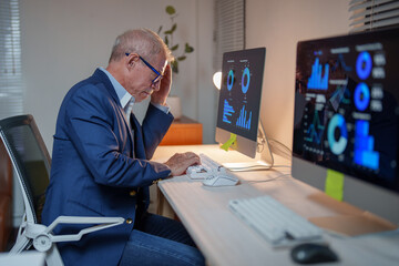 Senior businessman with glasses and blue jacket sitting at desk and looking at computer screen with declining financial charts, showing concern and touching his head with his hand