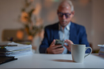 Focusing on a coffee cup in the foreground, a blurred businessman is using his smartphone in an office setting, surrounded by paperwork and a laptop on the desk