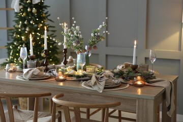Christmas table setting with stylish dishware, glasses and burning candles in festive decorated room