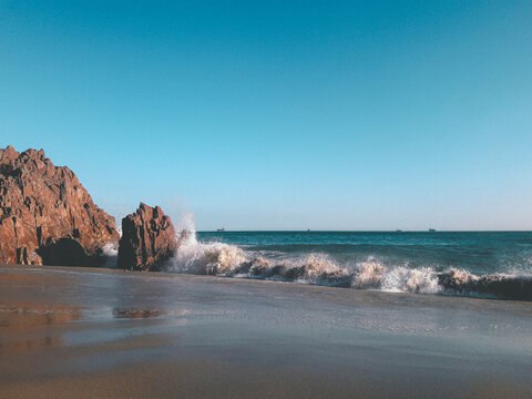 playa con arena y piedras rocosas grandes con olas en una tarde soleada y cielo azulado