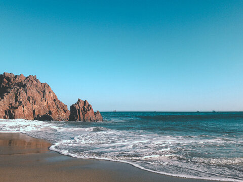 playa con arena y piedras rocosas grandes con olas en una tarde soleada y cielo azulado