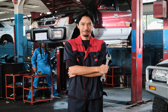 Portrait of Asian male professional mechanic with wrench, arms crossed stands in car fix garage, serious maintenance service jobs, and machine engineer work in automotive transport industry station.