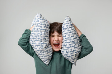 Annoyed boy covering his ears with pillows due to loud sound on grey background